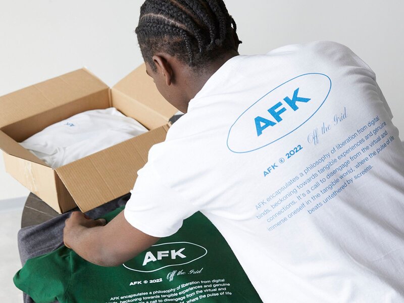 A man is unpacking a box containing t-shirts, with one shirt displayed on the table that has the text "AFK" printed on it.