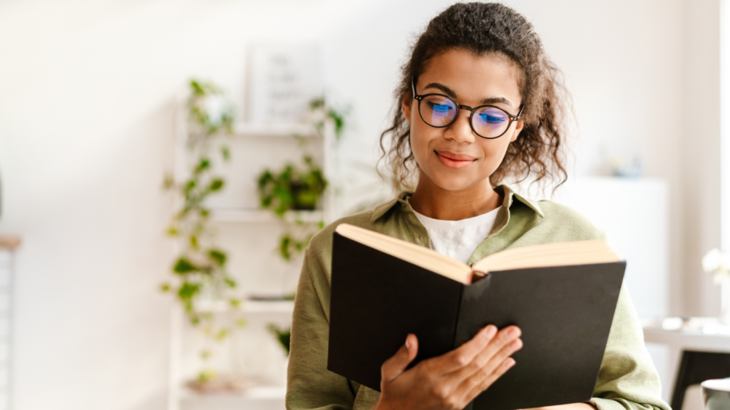 A young woman with glasses is intently reading a book, surrounded by a cozy, well-lit environment.