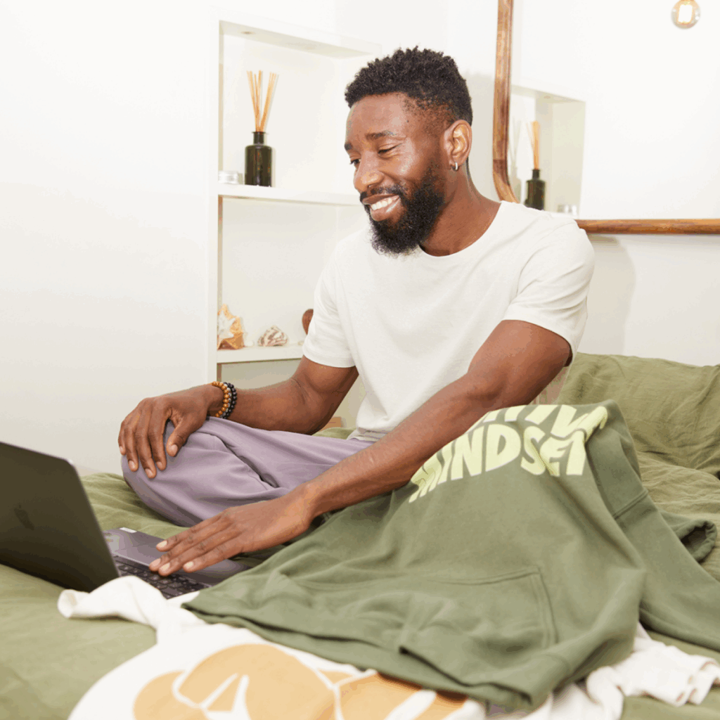 A smiling man is sitting on a bed with a laptop and a green hoodie with “Positive mindset” print nearby.