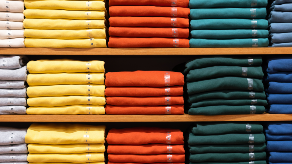 Stacks of neatly folded Gildan sweatshirts in yellow, orange, green, and blue are arranged on wooden shelves.