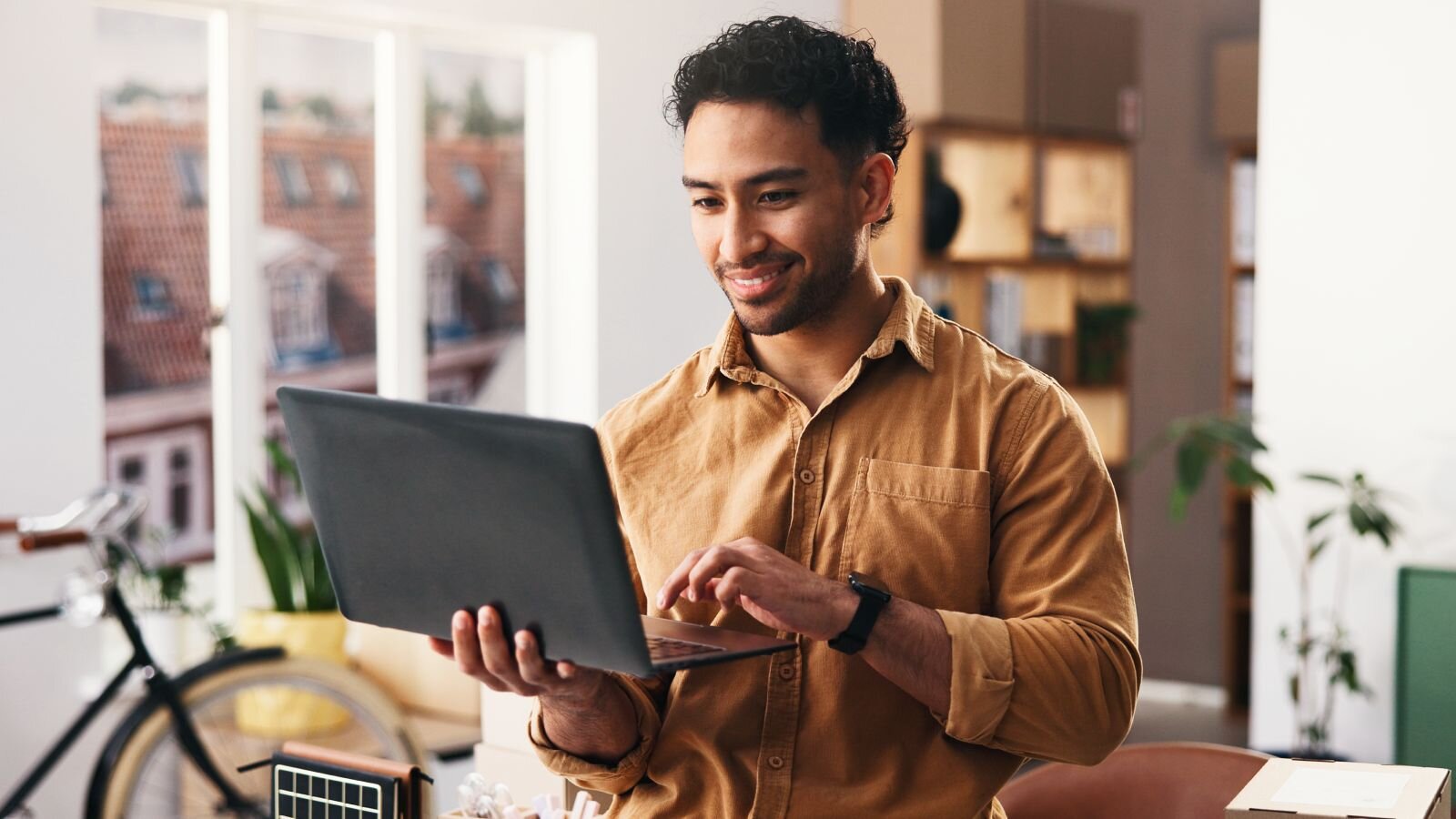 A smiling man in a brown shirt using a laptop in a bright, modern workspace with plants and a bicycle in the background.