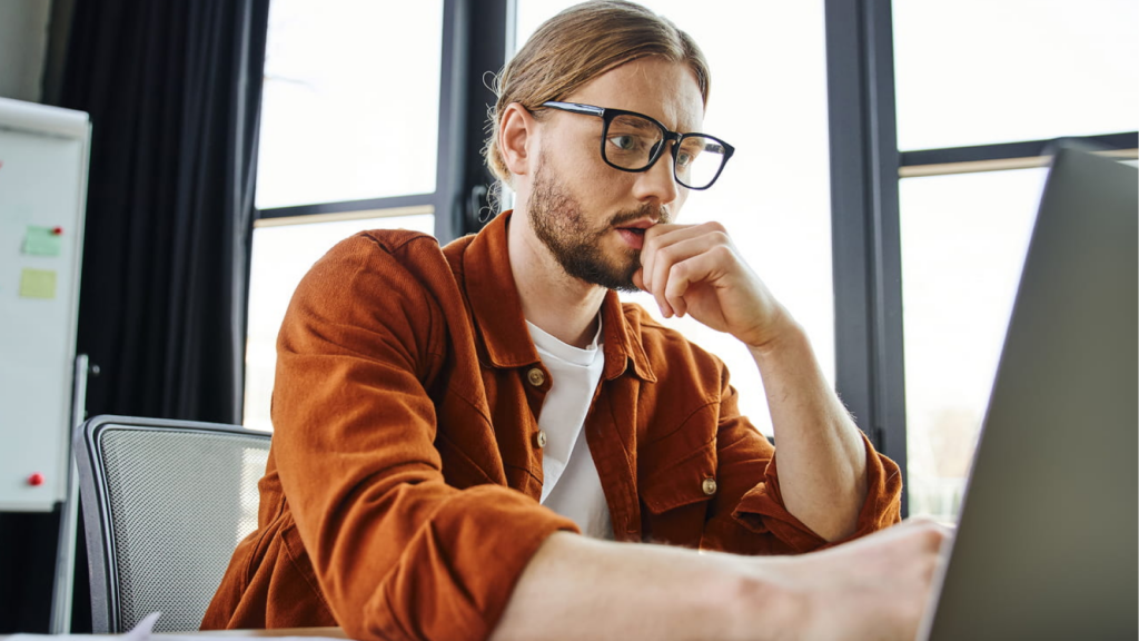 A man in a brown jacket, glasses, and a focused expression works on a laptop in an office.