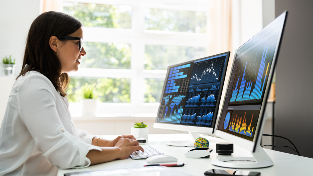 A woman seated at a desk, working on two computer monitors, focused on her tasks.