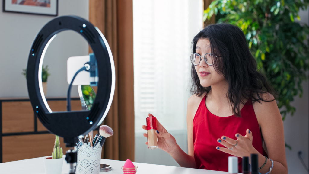 A woman in glasses presents make-up products during a livestream, using a smartphone on a stand with a ring light.