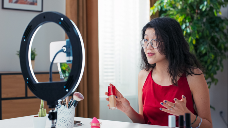 A woman in glasses presents make-up products during a livestream, using a smartphone on a stand with a ring light.
