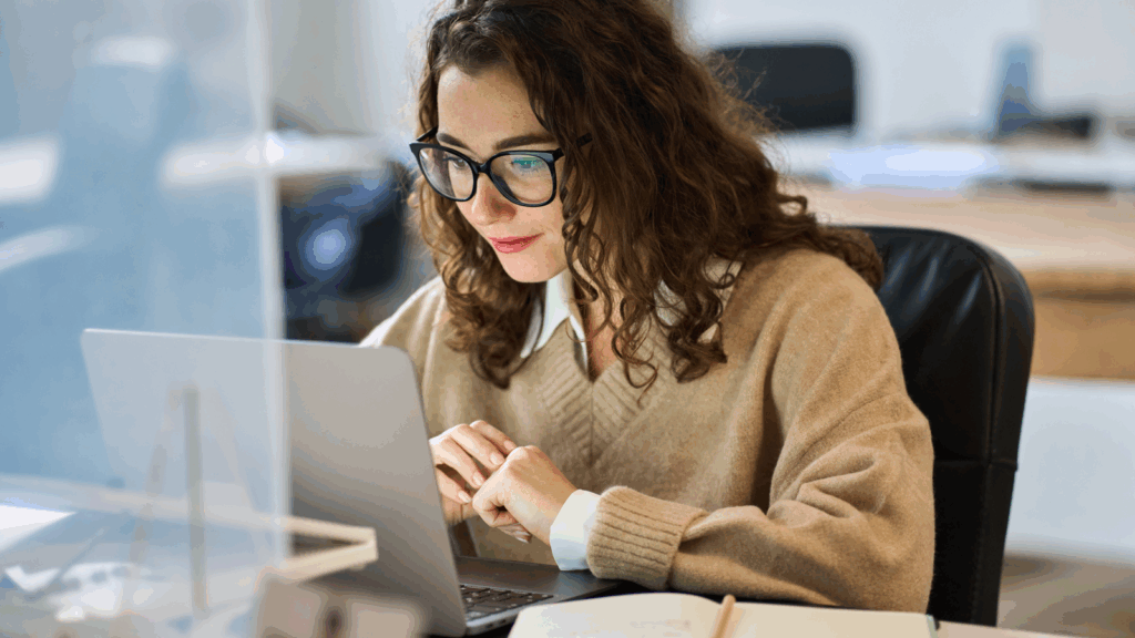 A woman wearing glasses is focused on her laptop, engaged in work at a desk.