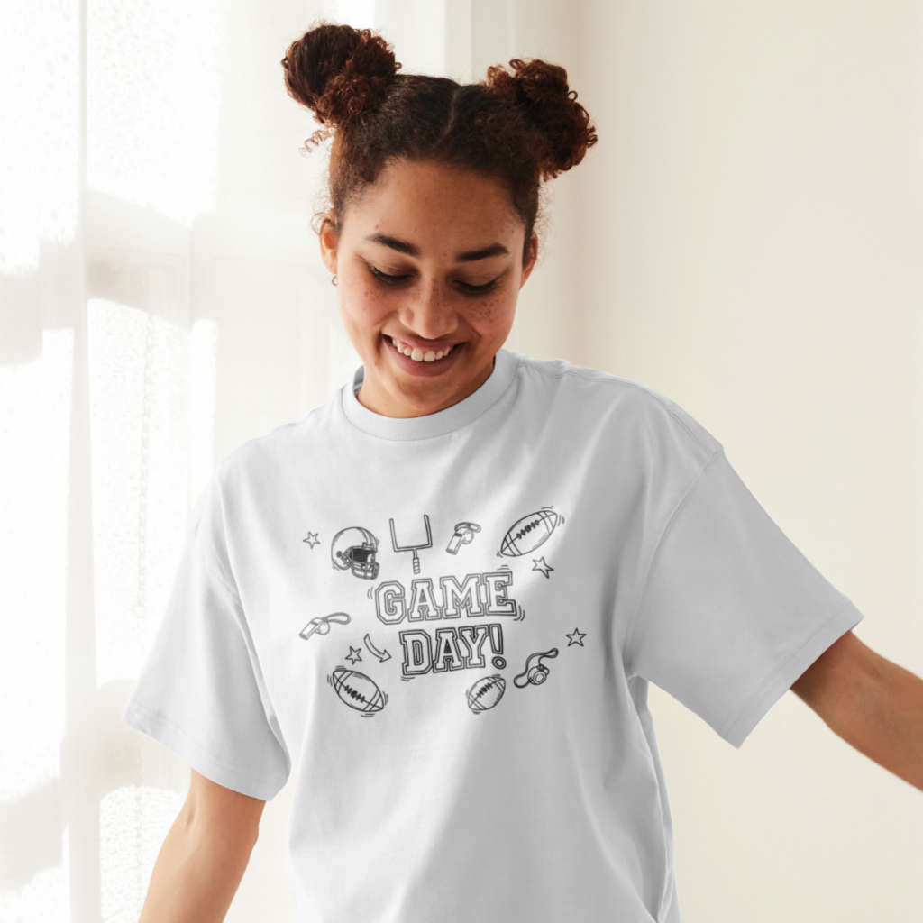A smiling woman wearing a white "Game Day" t-shirt with football elements drawn as doodles, standing by a sunlit window.