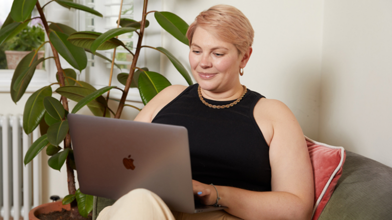 A smiling woman with short hair sits in a chair with a laptop, learning about the new Printify Pop-Up Store tools and features.