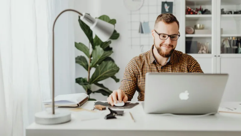 A smiling man sitting at a desk behind a laptop with his hand on the mouse.