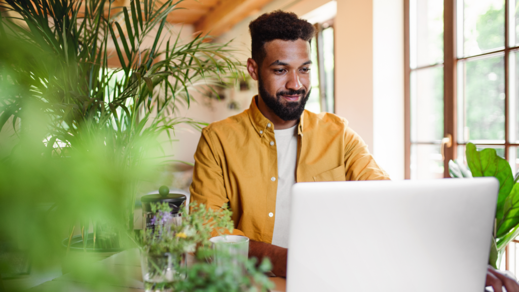 A man in a yellow shirt looking at a laptop screen and smiling while working.