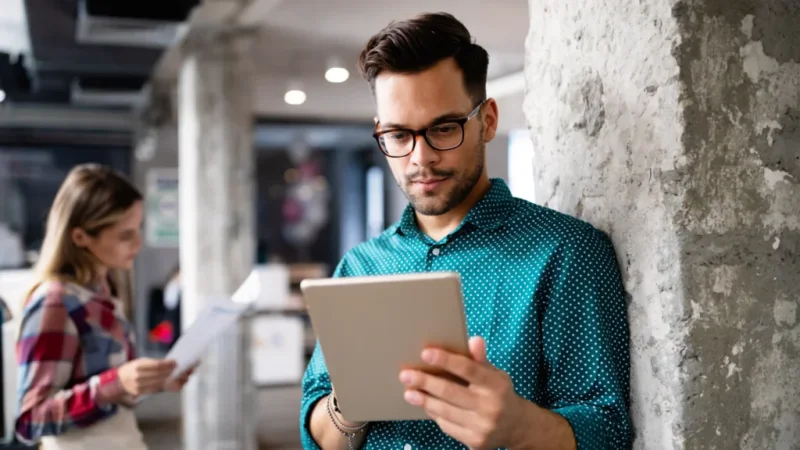 A man in glasses is leaning against a wall, reading from a tablet.
