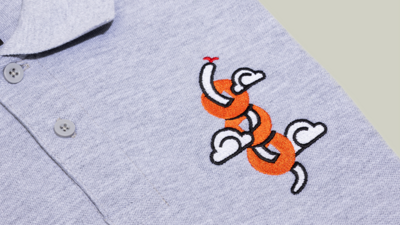 A close-up of a gray polo shirt featuring an embroidered design of intertwined shapes in orange and white.