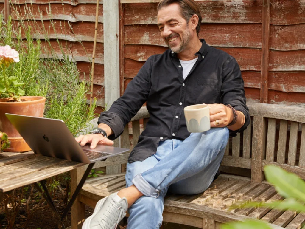 A man seated on a bench, working on a laptop and drinking tea or coffee
