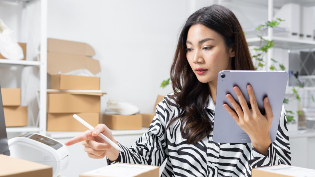 A woman holding a tablet in her hand and pointing with the other hand, with multiple delivery boxes in the background.