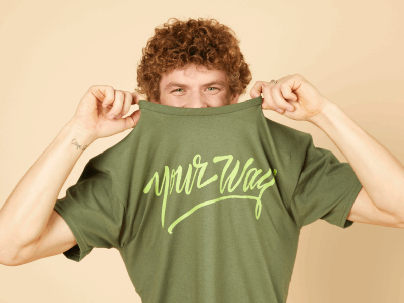 A man with curly hair playfully lifts a green t-shirt with "your way" text, partially covering his face.