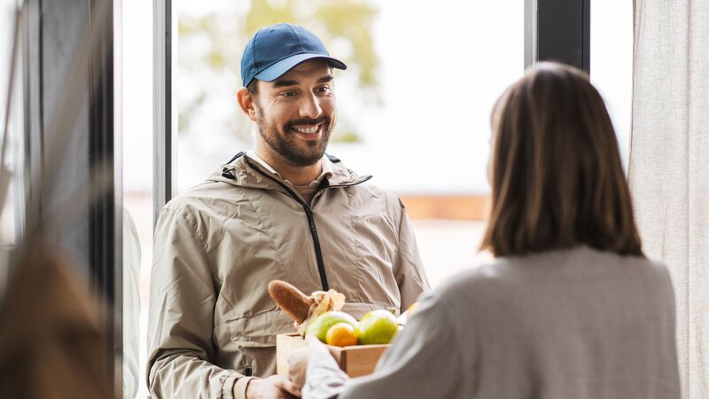 A smiling deliveryman in a blue cap hands a box of groceries, including apples and bread, to a woman at the door.