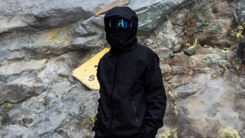 A person in visor glasses wearing a black gorpcore-themed suit. Large mountain rocks in the background.