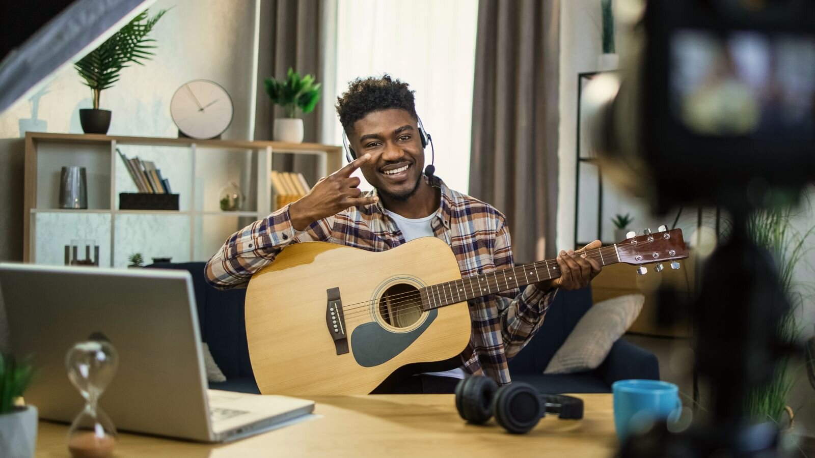 A smiling man wearing a flannel shirt is playing on a guitar and recording it using a camera.