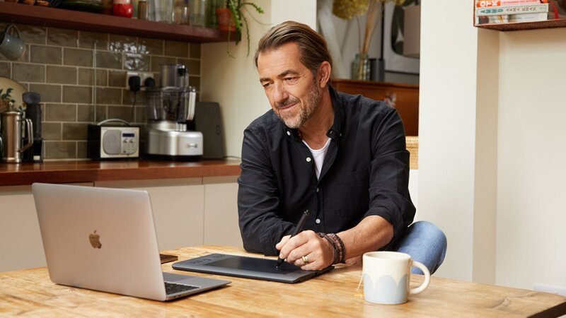 A man in a blue shirt and jeans is creating a text design with a pad and a laptop in a kitchen.