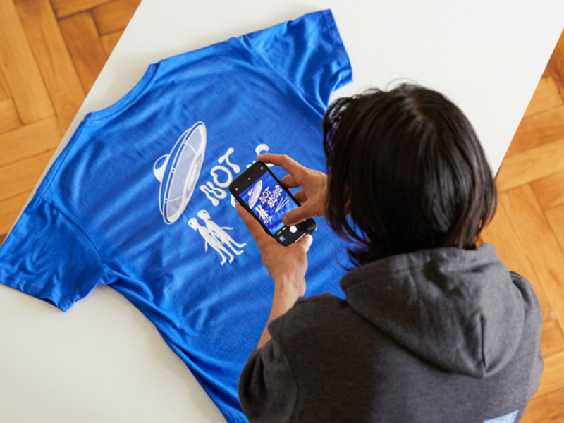 A young man with semi-long black hair in an asphalt grey hoodie is taking a picture of a blue “Not alone” alien-themed t-shirt.