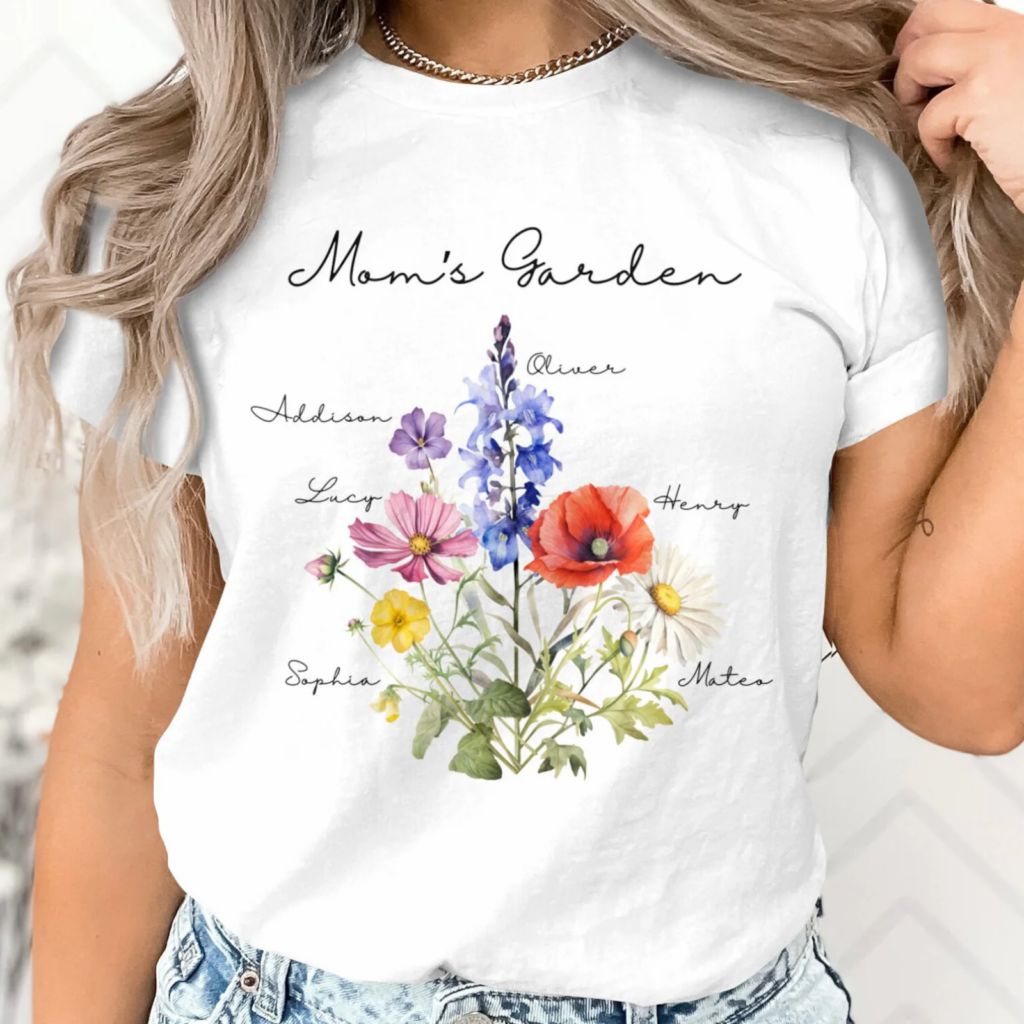 A woman wears a white t-shirt featuring colorful flowers labeled with names under the phrase "Mom's Garden".