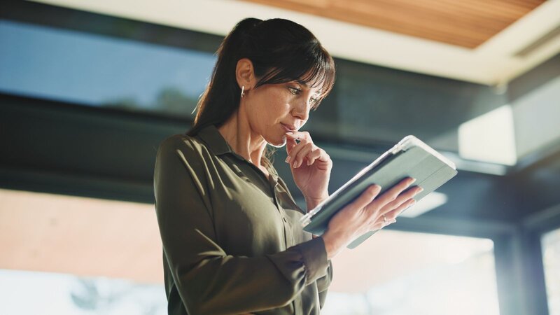 A woman standing indoors, looking thoughtfully at a tablet while pressing a finger against her lip.