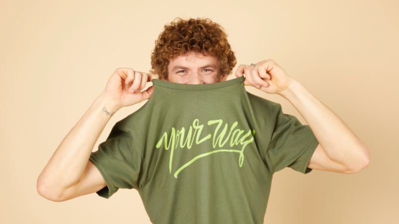 A young man with curly hair, wearing a green “Your way” t-shirt, pulls it halfway over his head. Light yellow background.