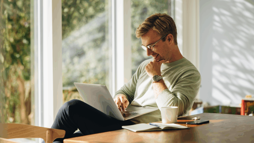 A man seated at a table, focused on his laptop, with a neutral background.