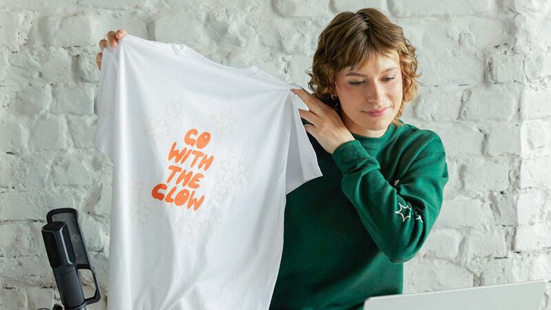 A young woman holding up a white t-shirt with the text "GO WITH THE GLOW" printed on it, sitting at a desk with a microphone and computer.