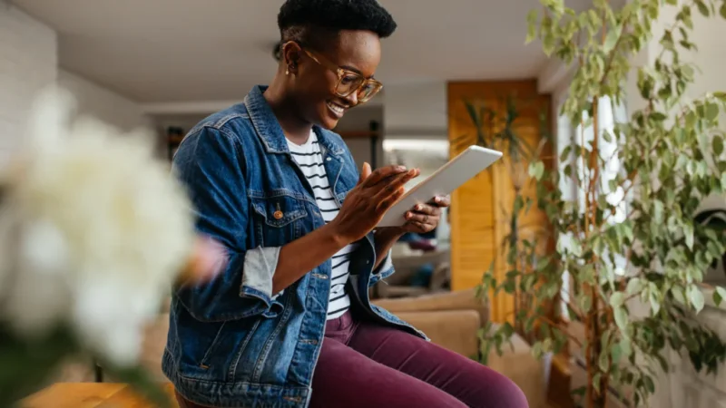 A woman in a denim jacket and glasses smiles while using a tablet for Etsy SEO. Seated in a cozy room with plants and soft lighting.