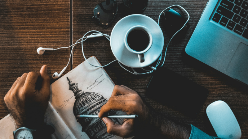 A person drawing a building in their notebook with a laptop in the background on the table.