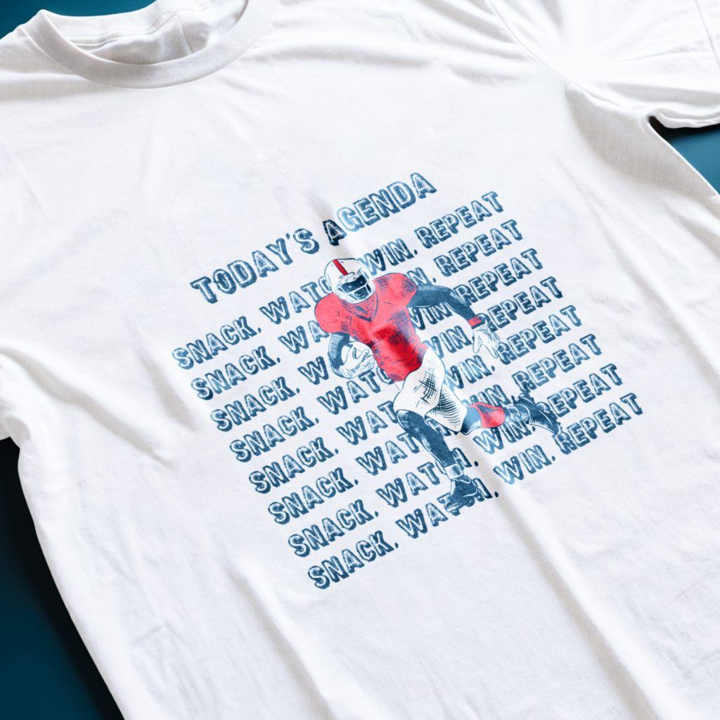 White t-shirt with a football player graphic in red and blue. Text says "Today's Agenda: Snack. Watch. Win. Repeat".