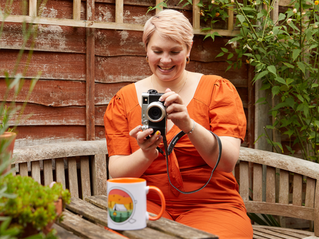 A smiling woman in an orange dress takes a picture of a colorful mug on the table, with greenery surrounding her.