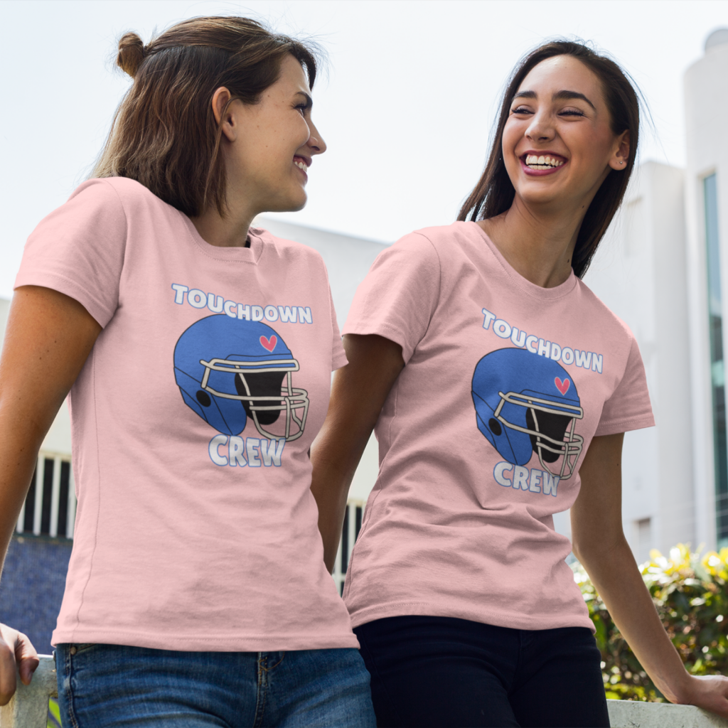 Two women smiling at each other, wearing matching pink t-shirts with a blue football helmet design and the words “Touchdown Crew”.