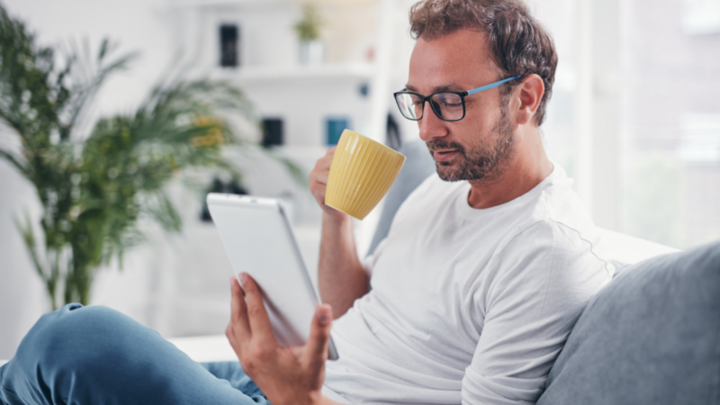A man in glasses is sitting on a sofa, holding a yellow coffee mug and reading a tablet for the best TikTok tools.