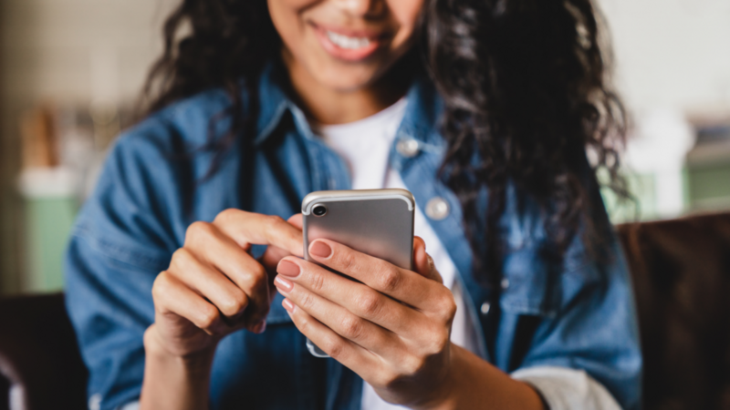 A woman in a blue denim shirt and a white t-shirt is holding a mobile phone in front of her. Focus on the phone, with the woman blurred.