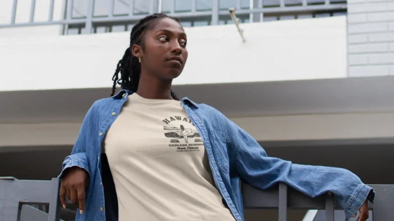 A woman is wearing a cream t-shirt with a left-chest logo, and a denim shirt. Urban scenery in the background.