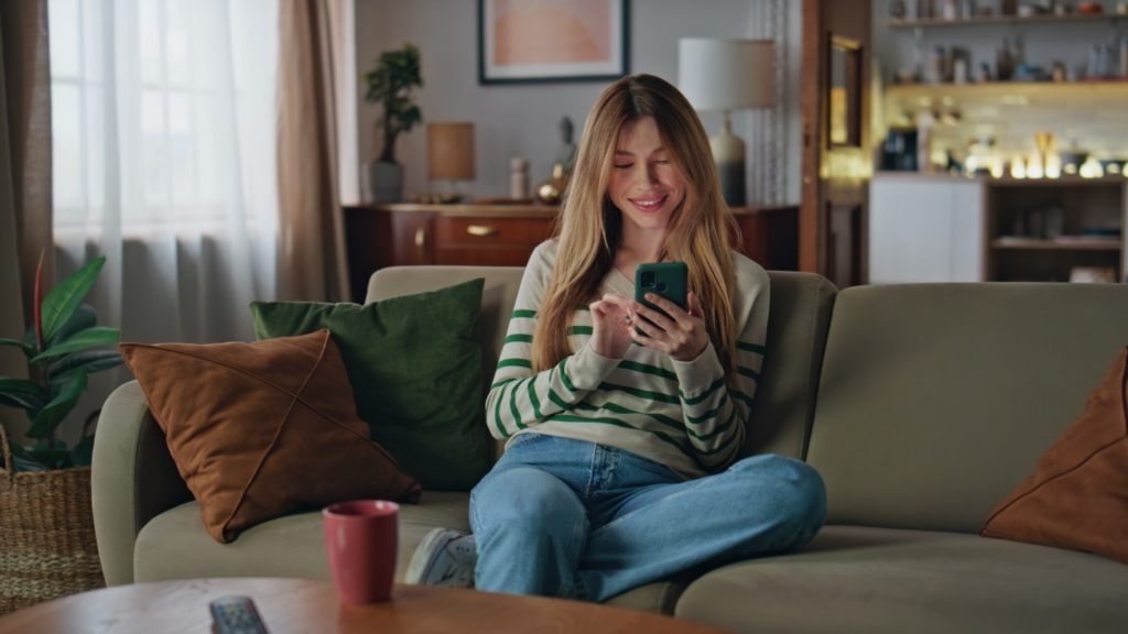 A woman in a green shirt and jeans sits on a yellow couch, using a smartphone, looking for the latest TikTok trends.