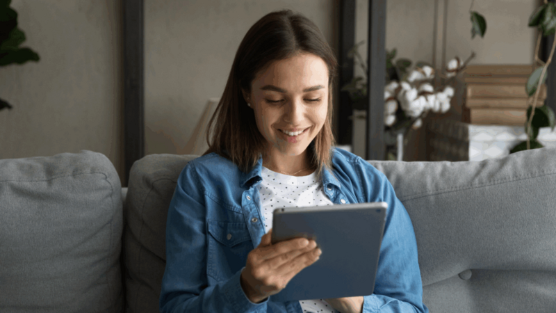 A smiling woman is sitting on a couch and using a tablet to find the best AI tools for eCommerce.