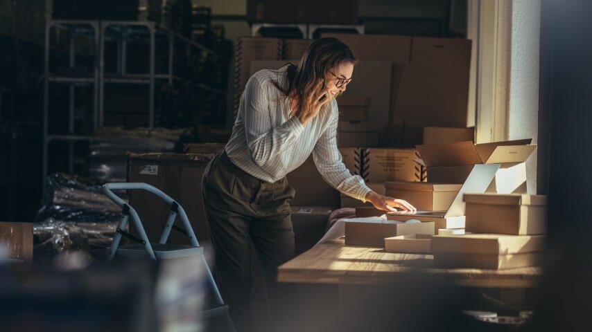 A woman talking on the phone, typing in laptop, boxes around her.