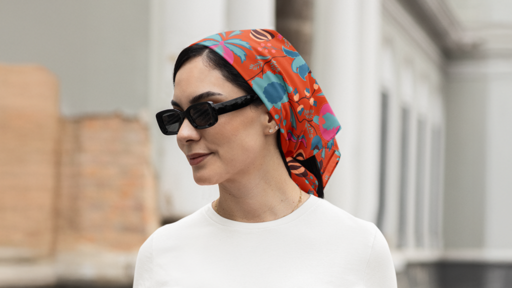 A woman in sunglasses and a vibrant red headscarf with floral patterns stands outdoors against a blurred background, wearing a white top.