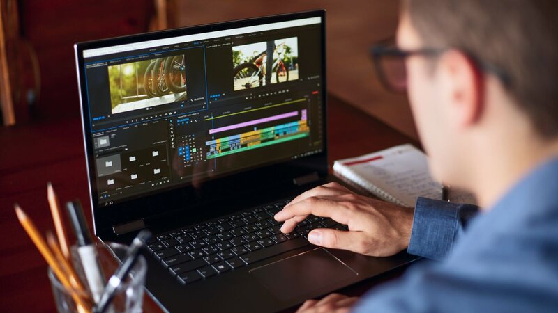 A person edits a video on a laptop, with colorful timelines and clips visible on the screen. The desk has pencils and a notebook.