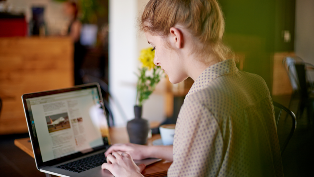 A woman with blonde hair works on a laptop, comparing Shopify vs Etsy, in a cozy cafe, focused on the screen. Yellow flowers in a vase nearby.