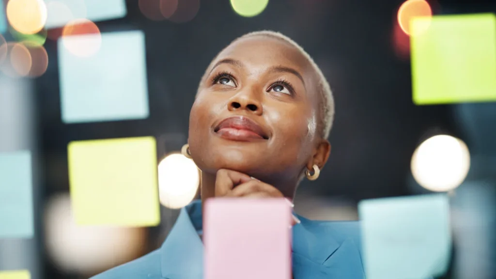 A woman in a business suit gazes thoughtfully at a colorful post-it note board filled with notes and ideas.