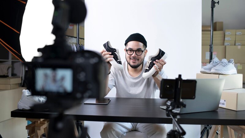 A man in a white t-shirt and a black beanie is holding two pairs of shoes, sitting at a table, while being filmed on camera.