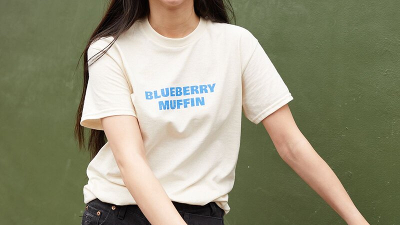 A person wearing a beige t-shirt with "BLUEBERRY MUFFIN" printed in blue letters.