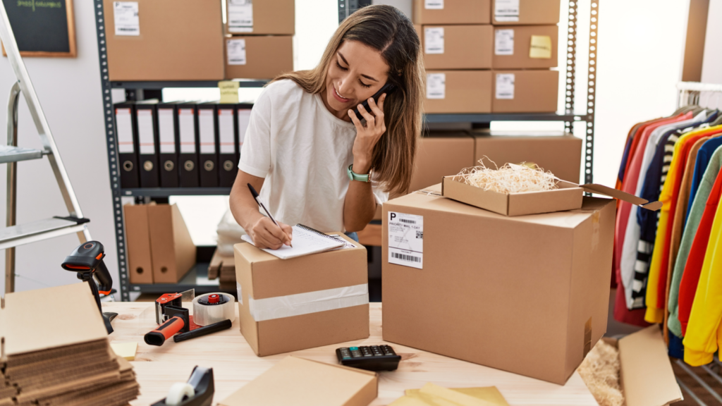 A woman sits at a desk, focused on her computer, surrounded by boxes and papers, engaged in her work.