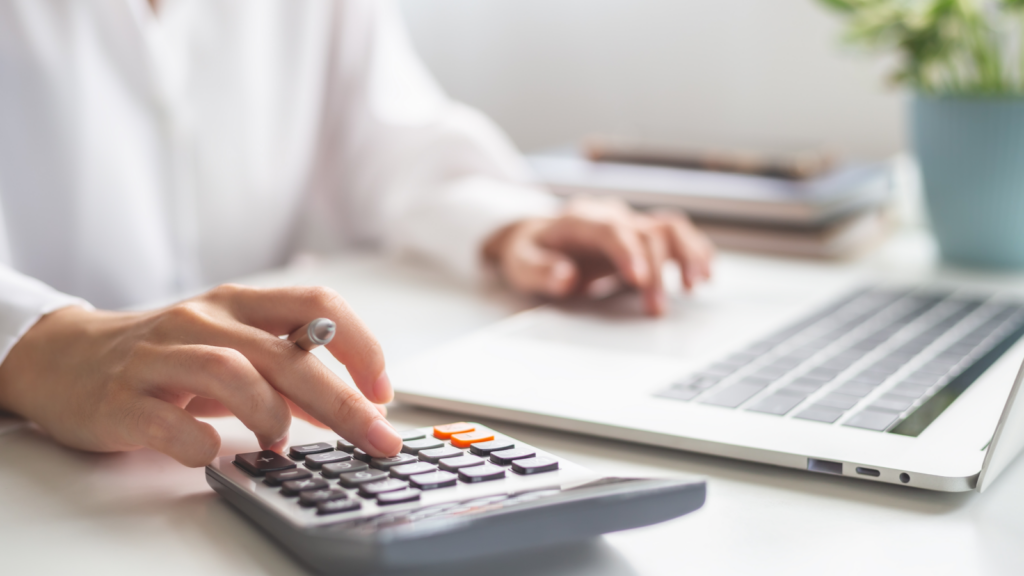 A woman sitting at an office desk with a laptop on top of it and using a calculator.
