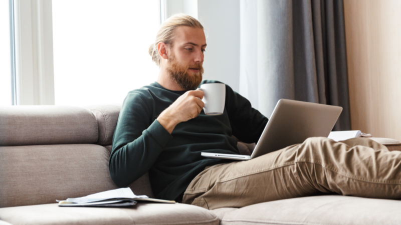 A bearded man is sitting on a couch with his legs up, drinking coffee and looking at a laptop.