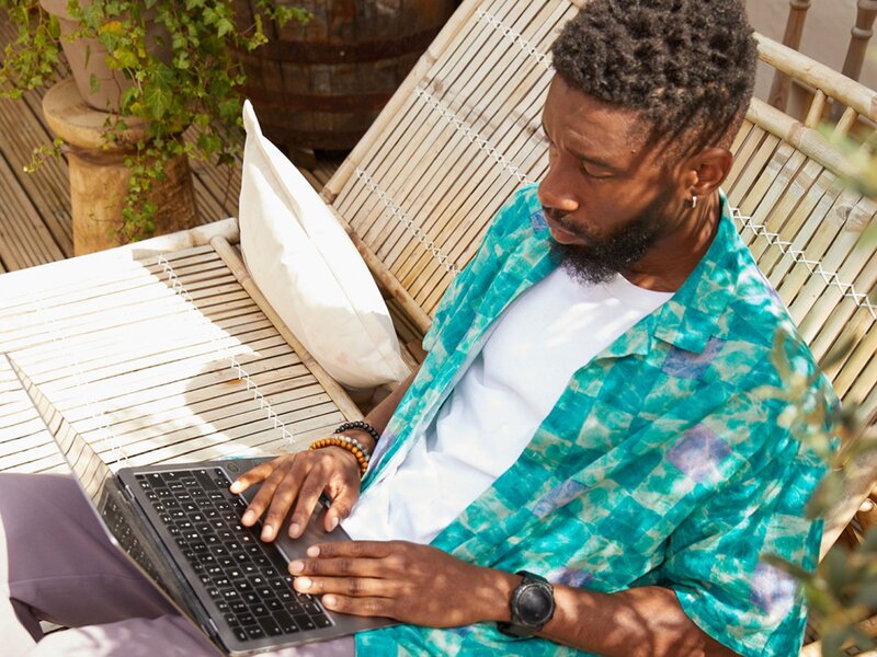 A bearded man is sitting outside on a wooden bench, using a laptop and wearing a colorful blue shirt.